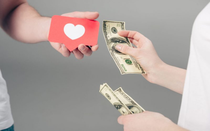 cropped view of woman giving dollar banknotes to man with red paper cut card with heart symbol on